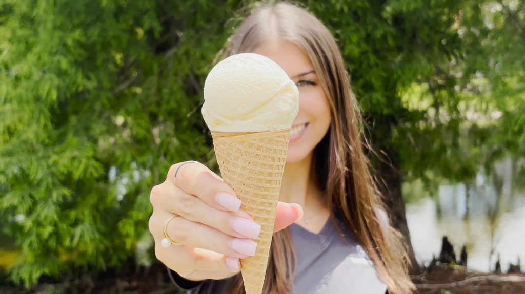 A smiling girl holding up a cone of vanilla ice cream.